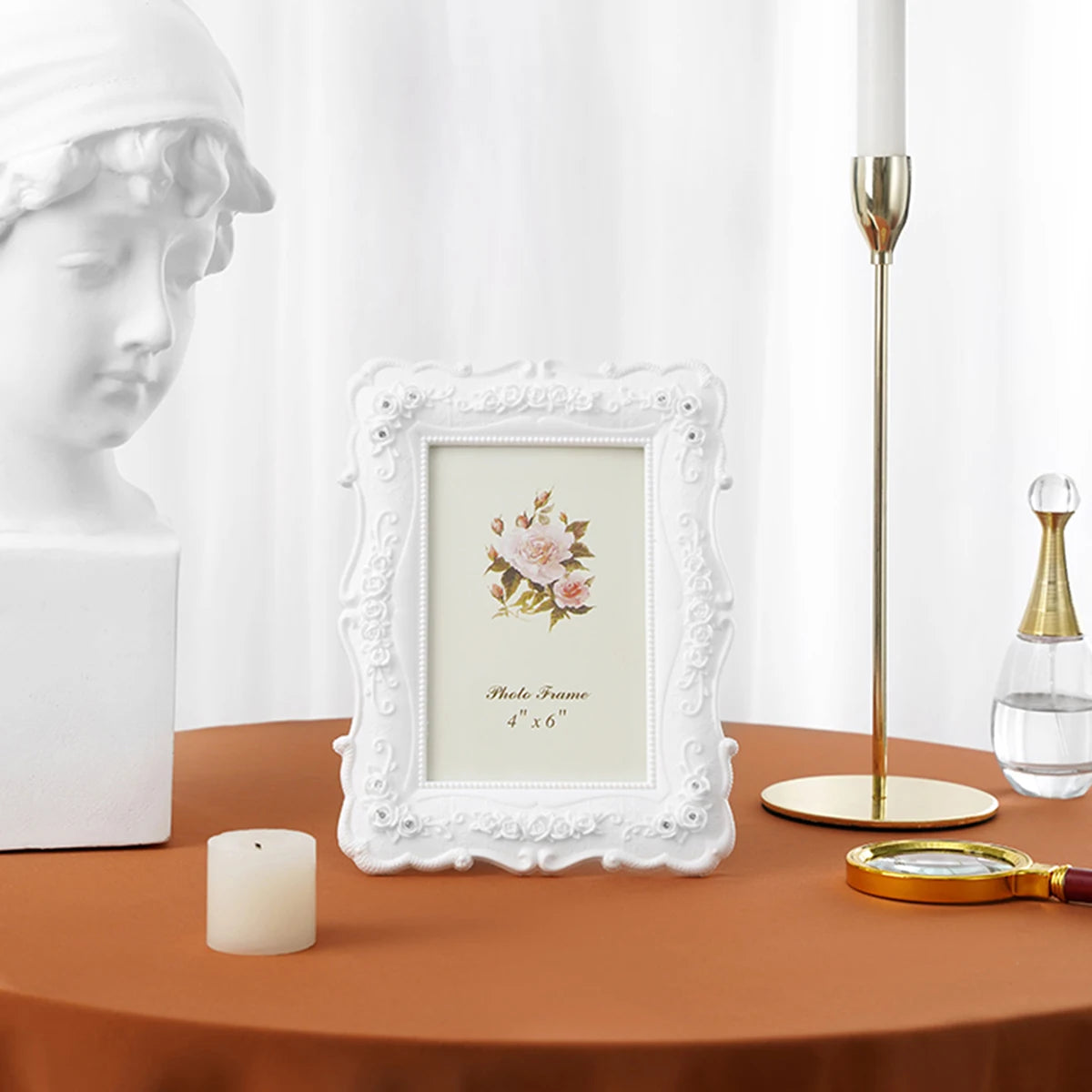 A white ornate photo frame with a pink rose design sits on a brown table next to a white statue and a gold candle holder.