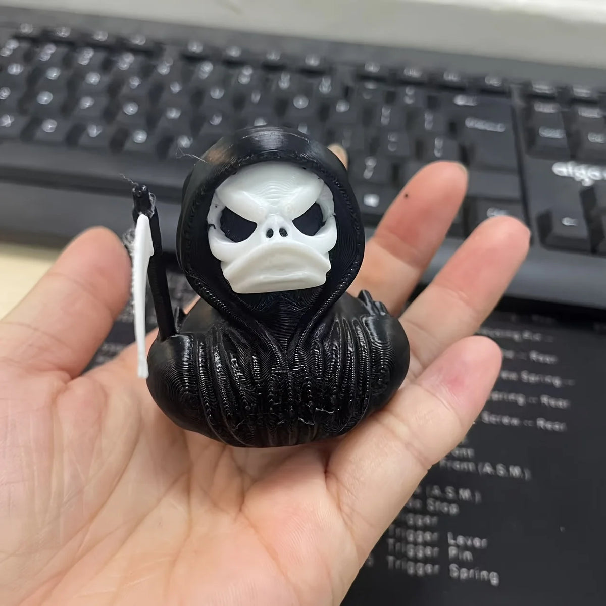 A black 3D printed statue of a Grim Reaper duck with a skull hood is held in a hand.