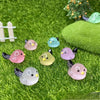 A set of small, luminous resin bird fairy garden decor featuring pink, green, and yellow birds on green moss.