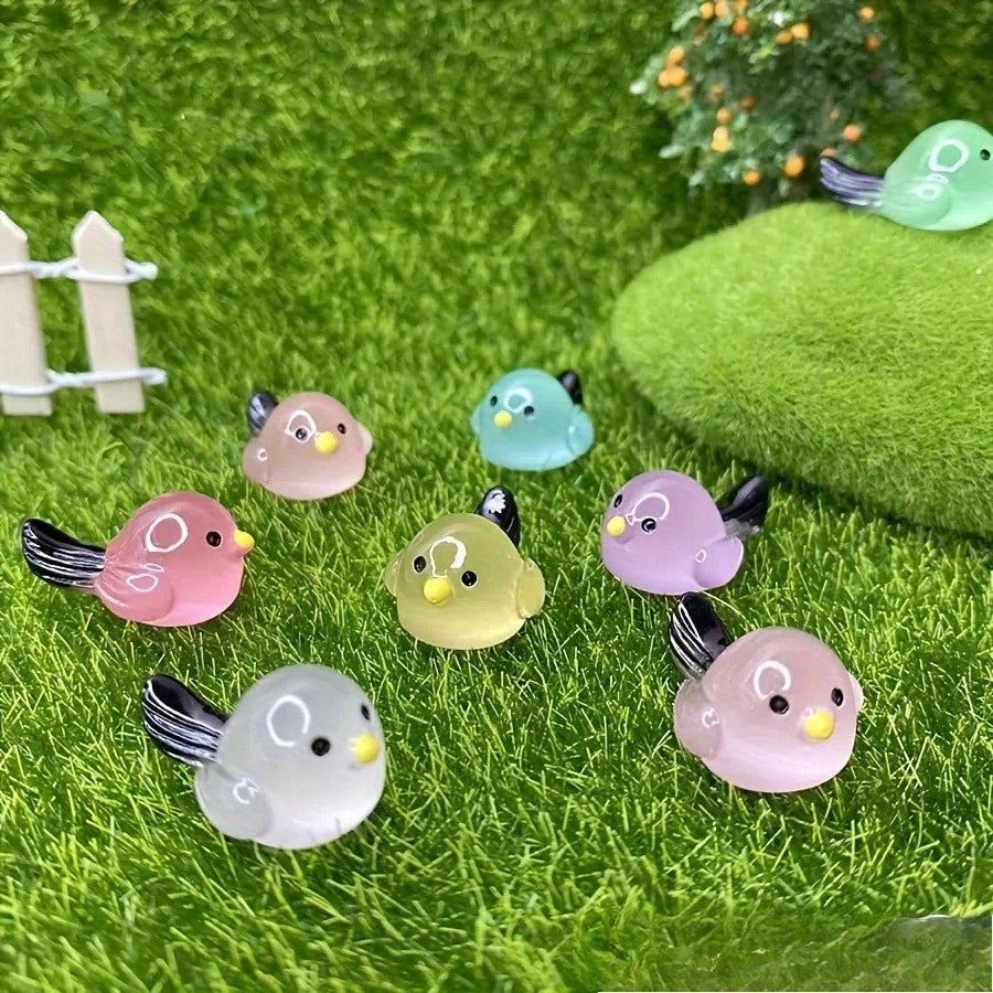 A set of small, luminous resin bird fairy garden decor featuring pink, green, and yellow birds on green moss.