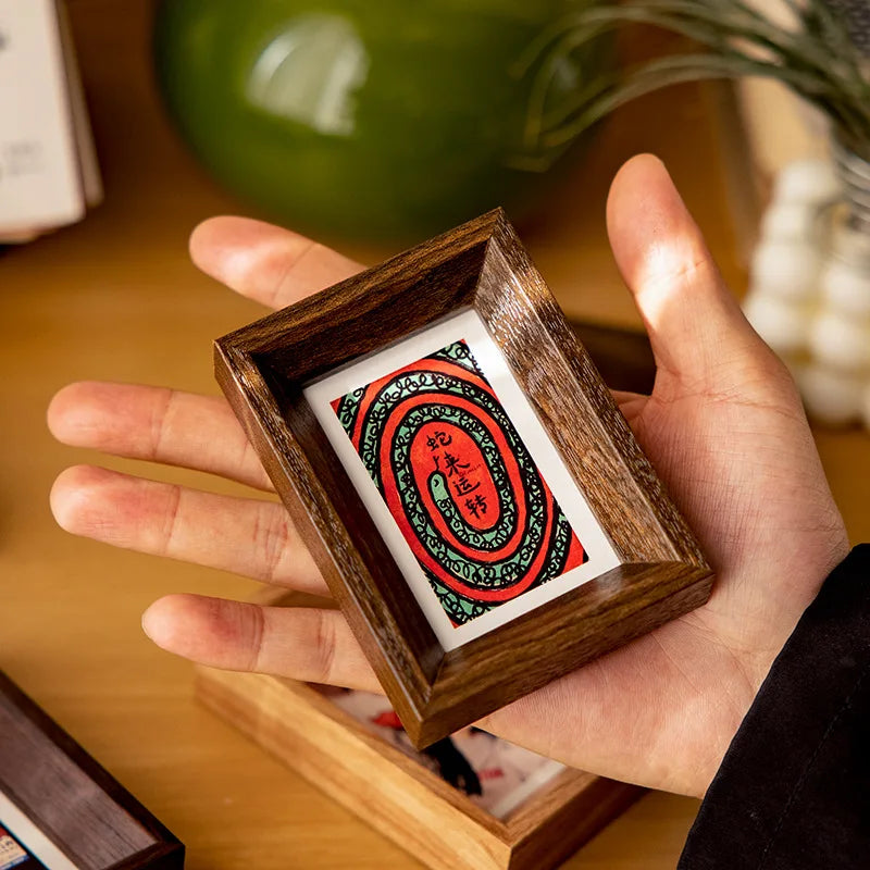 A hand holds a small wooden photo frame containing an orange and green patterned card.
