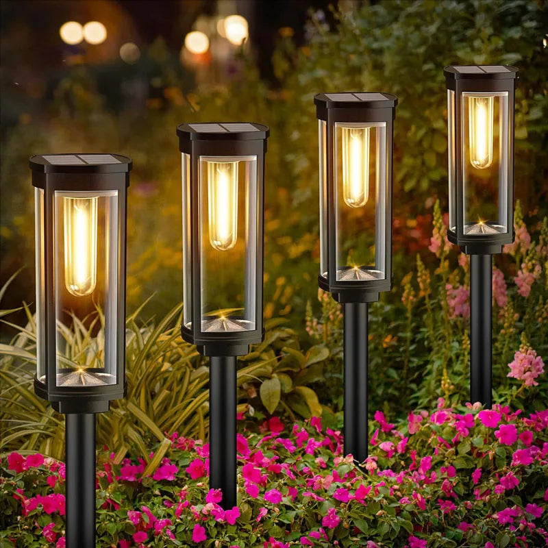 A row of black retro solar pathway lights illuminates a garden bed with pink flowers at night.