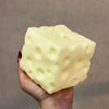 A hand holds a large, pale yellow cheese cube stress ball with visible holes and dimples.