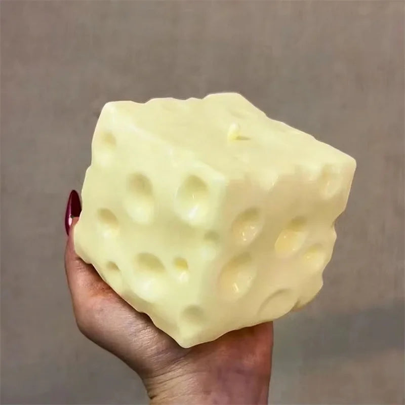 A hand holds a large, pale yellow cheese cube stress ball with visible holes and dimples.