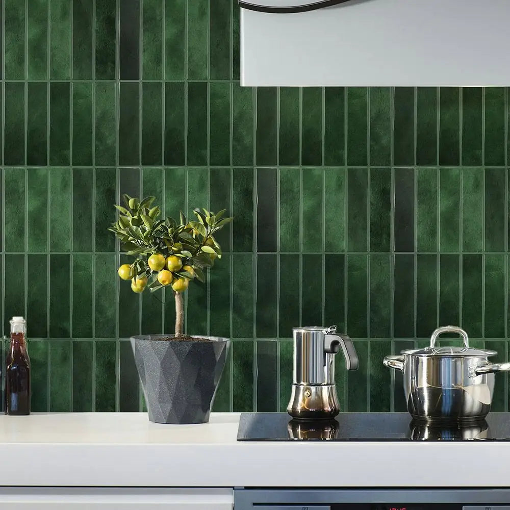 A kitchen countertop with a dark green tiled backsplash featuring a potted olive tree, a stainless steel kettle, and a silver
