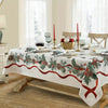 A white Christmas themed tablecloth with red and green foliage and ribbon on a wooden table.