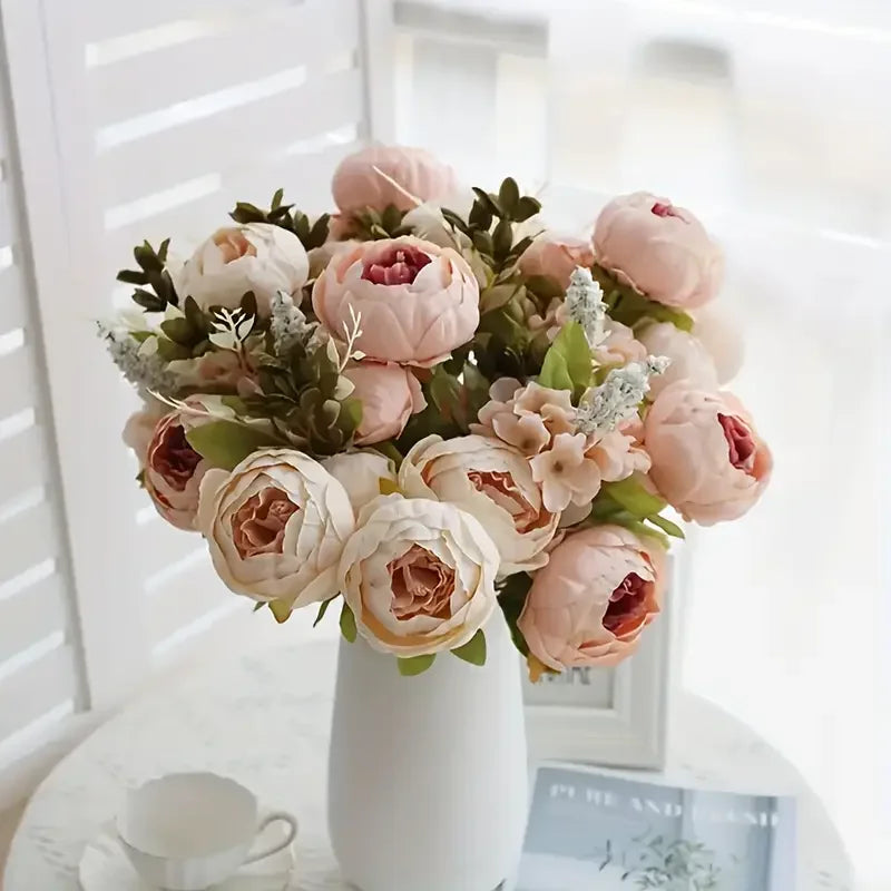 A bouquet of pink and peach peony artificial flowers with green foliage in a white vase.