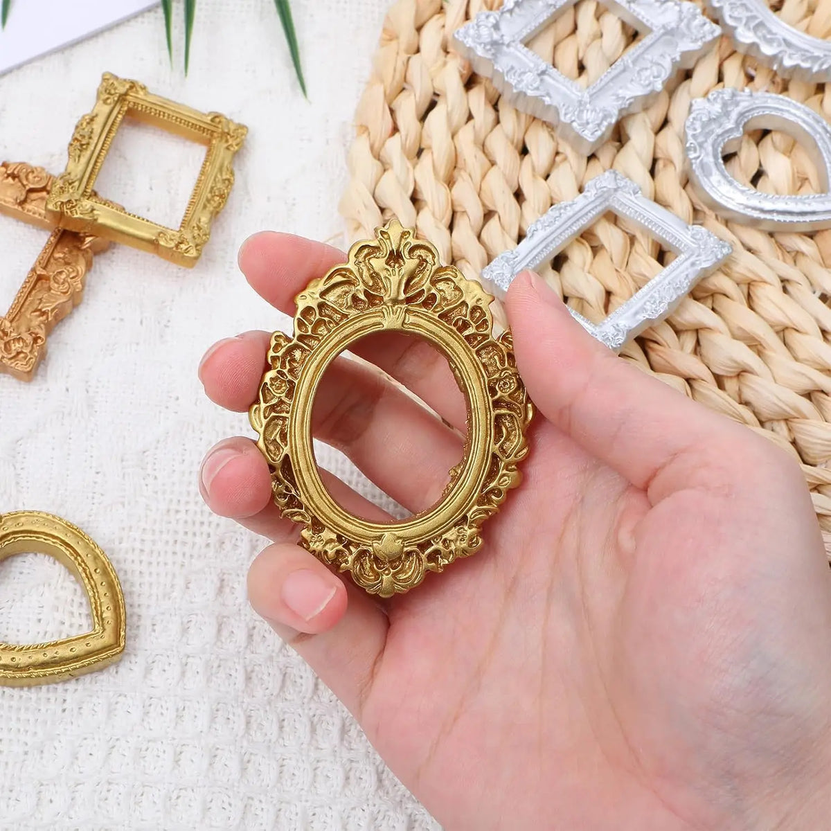 A hand holds a vintage gold mini picture frame surrounded by silver and gold frames on a woven mat.