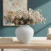 A rustic textured round white vase filled with dried floral arrangements sits on a wooden table against a teal wall.