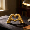 A golden heart shape sculpture made of two hands resting together on a dark surface.