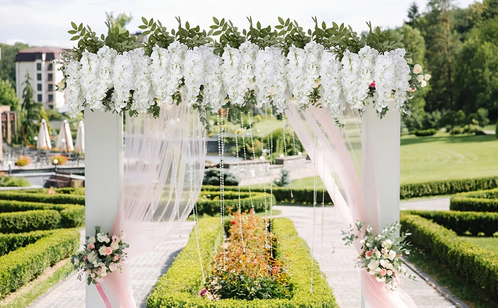 A white floral archway decorated with artificial wisteria vines and silk bloom forks in a garden setting.