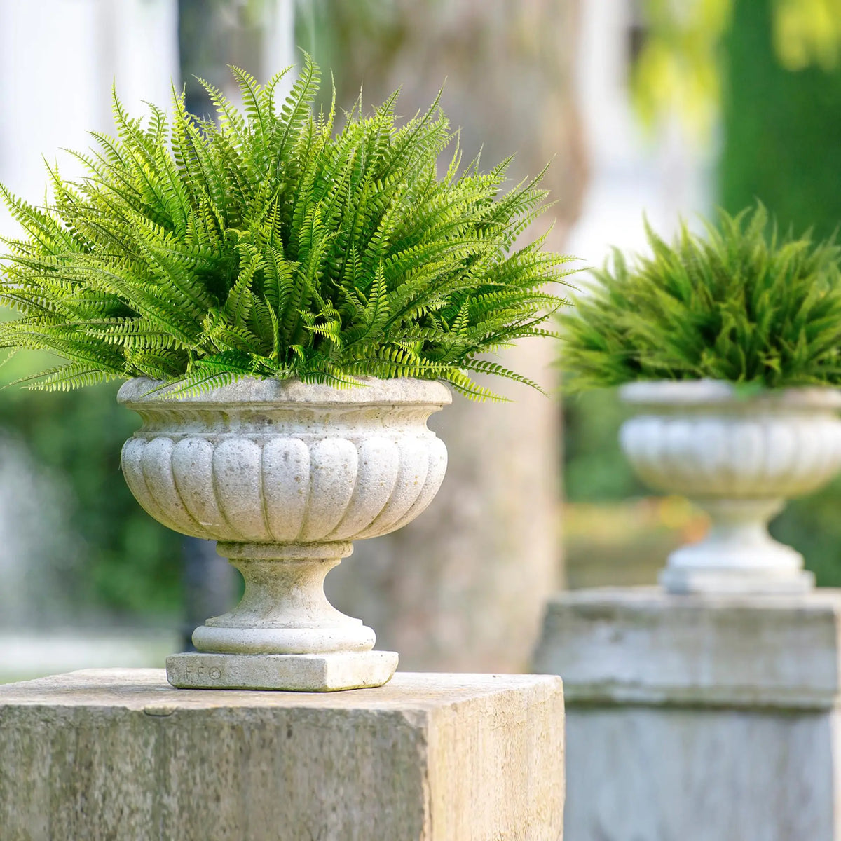 A potted artificial Boston fern in a stone urn sits on an outdoor surface with blurred greenery in the background.