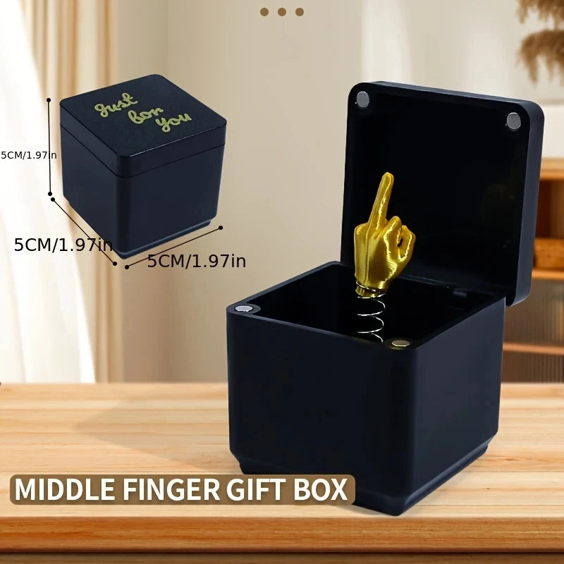 Boîte cadeau avec un doigt d'honneur noir et un geste de main doré, sur une surface en bois.