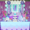 A table set for a mermaid party featuring purple and teal plates, cups, and napkins decorated with purple tulle and pink acce