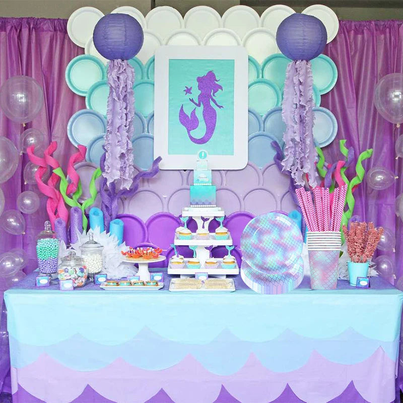 A table set for a mermaid party featuring purple and teal plates, cups, and napkins decorated with purple tulle and pink acce