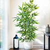 Artificial bamboo branch with green leaves displayed in a white pot next to a window.