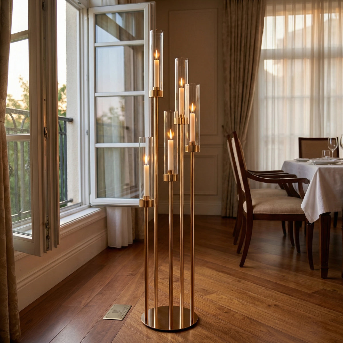 A tall, slender gold LED candle holder with five arms stands on a wooden floor next to a window.