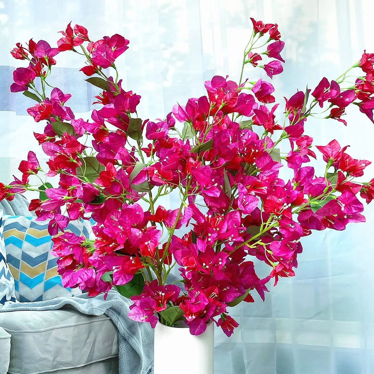 Leuchtend magentafarbene künstliche Bougainvillea-Blüten in einem weißen Topf vor blauem Hintergrund.