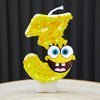 A yellow sponge cake candle shaped like the number 3, decorated with yellow glitter and a face.
