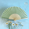 A light green folding fan with delicate perforations and a glass chain accessory is displayed against a blue background.