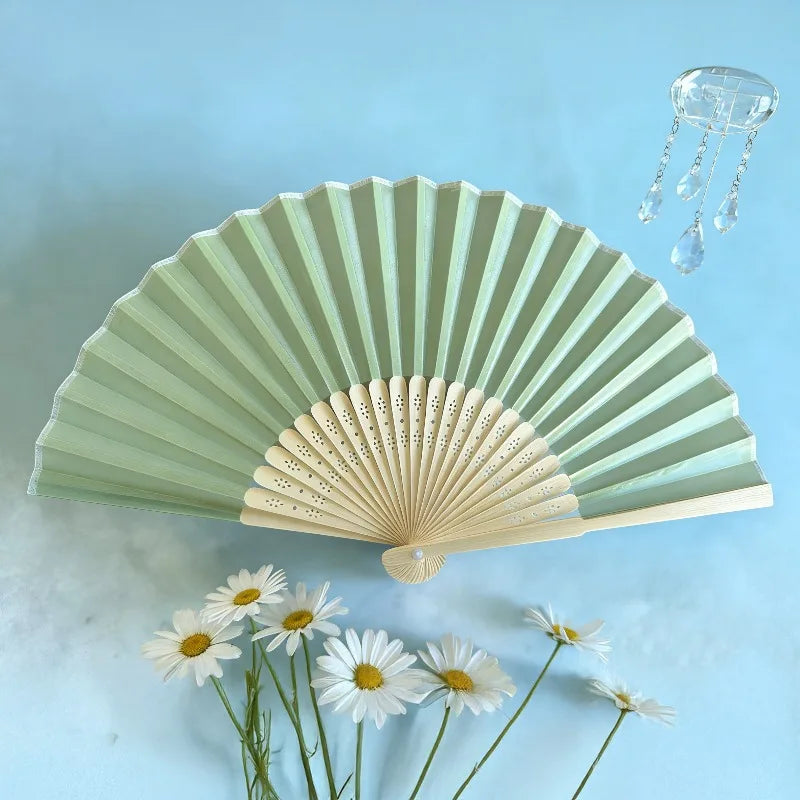 A light green folding fan with delicate perforations and a glass chain accessory is displayed against a blue background.