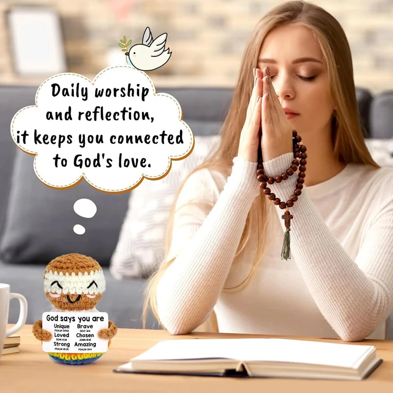 A woman with long hair is praying, holding hands together, next to a crochet doll and an open book.