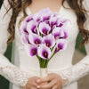 A bouquet of six artificial purple calla lilies held by a person wearing a white dress.