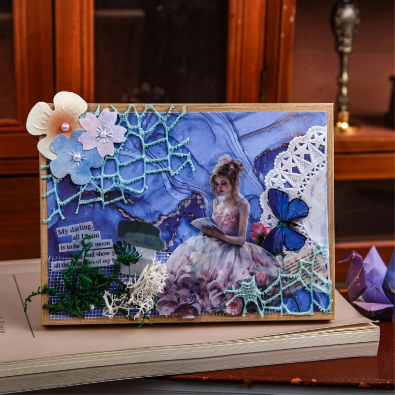 A handmade paper art piece featuring a watercolor floral design with blue and purple tones and white lace accents.