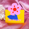 A bright yellow card featuring a pink flower design and the text Be Happy, placed on a pink fabric.