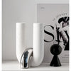 A white ceramic U-shaped tabletop vase with a silver metallic accent sits on a white surface next to a black spherical object