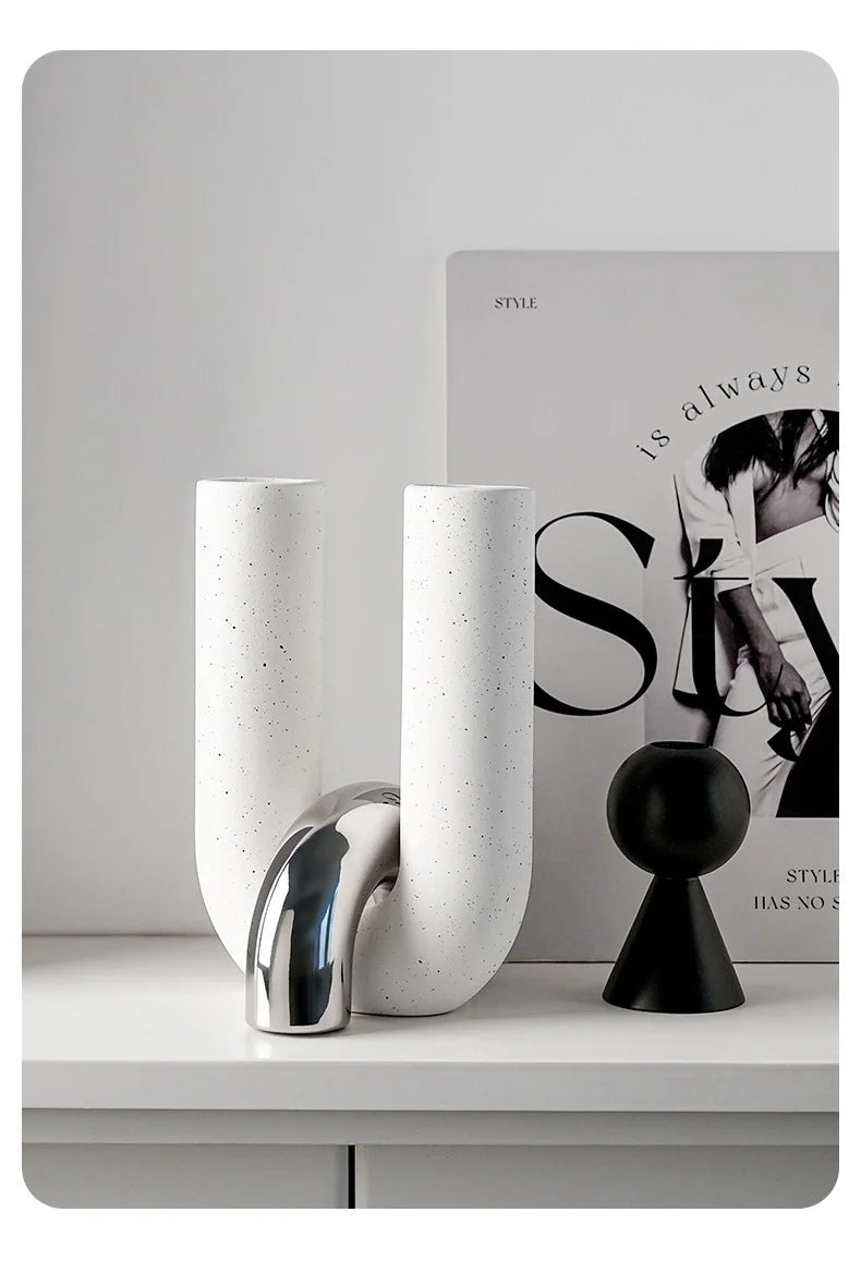 A white ceramic U-shaped tabletop vase with a silver metallic accent sits on a white surface next to a black spherical object
