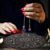 A hand holds a silver chain pendant with a purple gemstone over a wooden divination board.