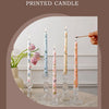 A collection of tall, slender printed candles in pink and green floral patterns displayed on a light surface.