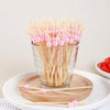 A glass filled with bamboo fruit skewers featuring pink and white spherical beads resting on a wooden board.