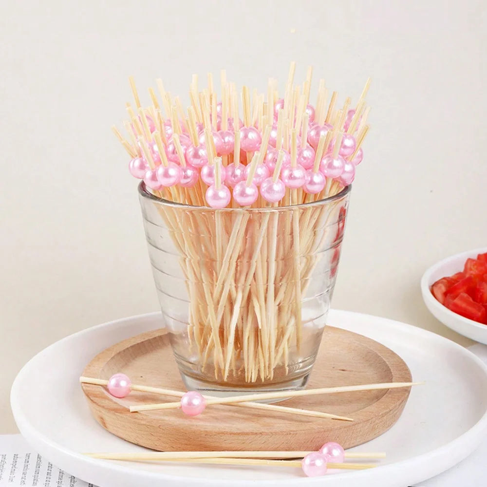 A glass filled with bamboo fruit skewers featuring pink and white spherical beads resting on a wooden board.