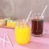 A glass jar of orange juice with a straw next to a glass of iced tea and several clear disposable drinking straws on a pink s