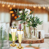 A glass vase holds greenery and white flowers next to elegant gold-rimmed champagne flutes on a wooden slab.