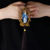 A pair of hands holds a wooden pendant ornament depicting Our Lady of Grace with a halo and golden wreath.