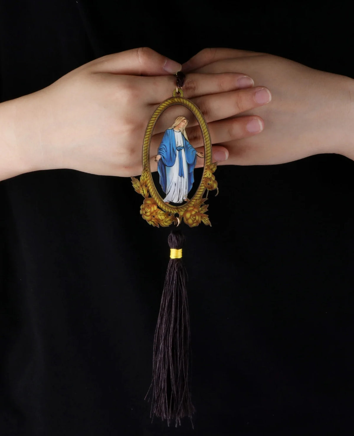 A pair of hands holds a wooden pendant ornament depicting Our Lady of Grace with a halo and golden wreath.