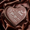 A clear acrylic heart plaque engraved with 'I love you dear Dad' resting on dark brown satin fabric.