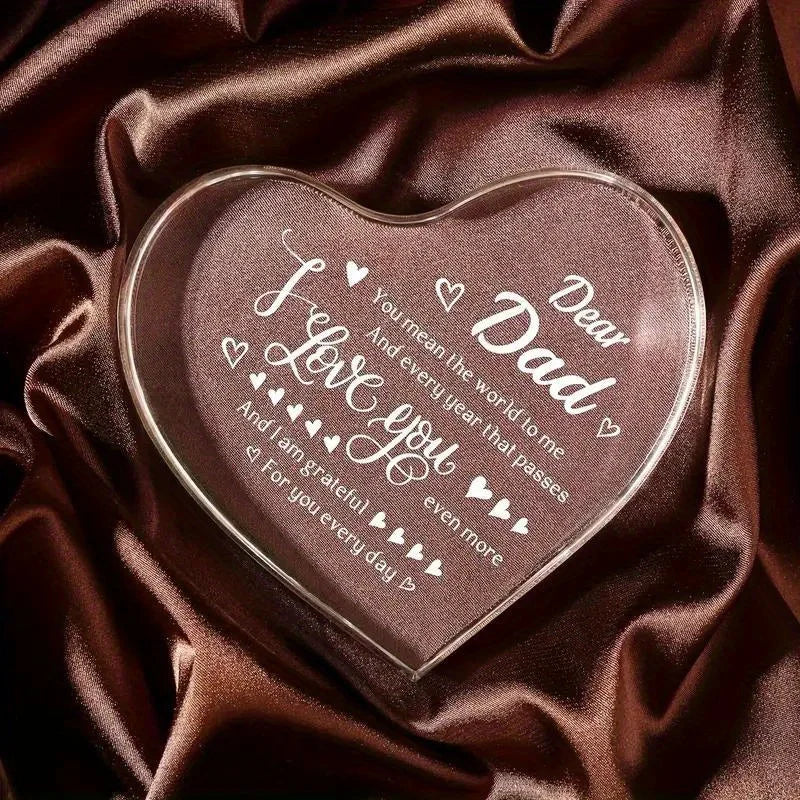 A clear acrylic heart plaque engraved with 'I love you dear Dad' resting on dark brown satin fabric.
