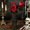 A dark gothic decorative vase shaped like a skull holding a bouquet of red roses and lit candles.