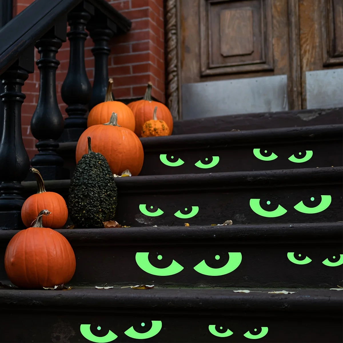 A set of eight glow-in-the-dark Halloween eye window clings are placed on a dark staircase next to orange pumpkins.