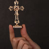 A hand holds a gold metal crucifix desk cross with a self-adhesive base against a dark background.