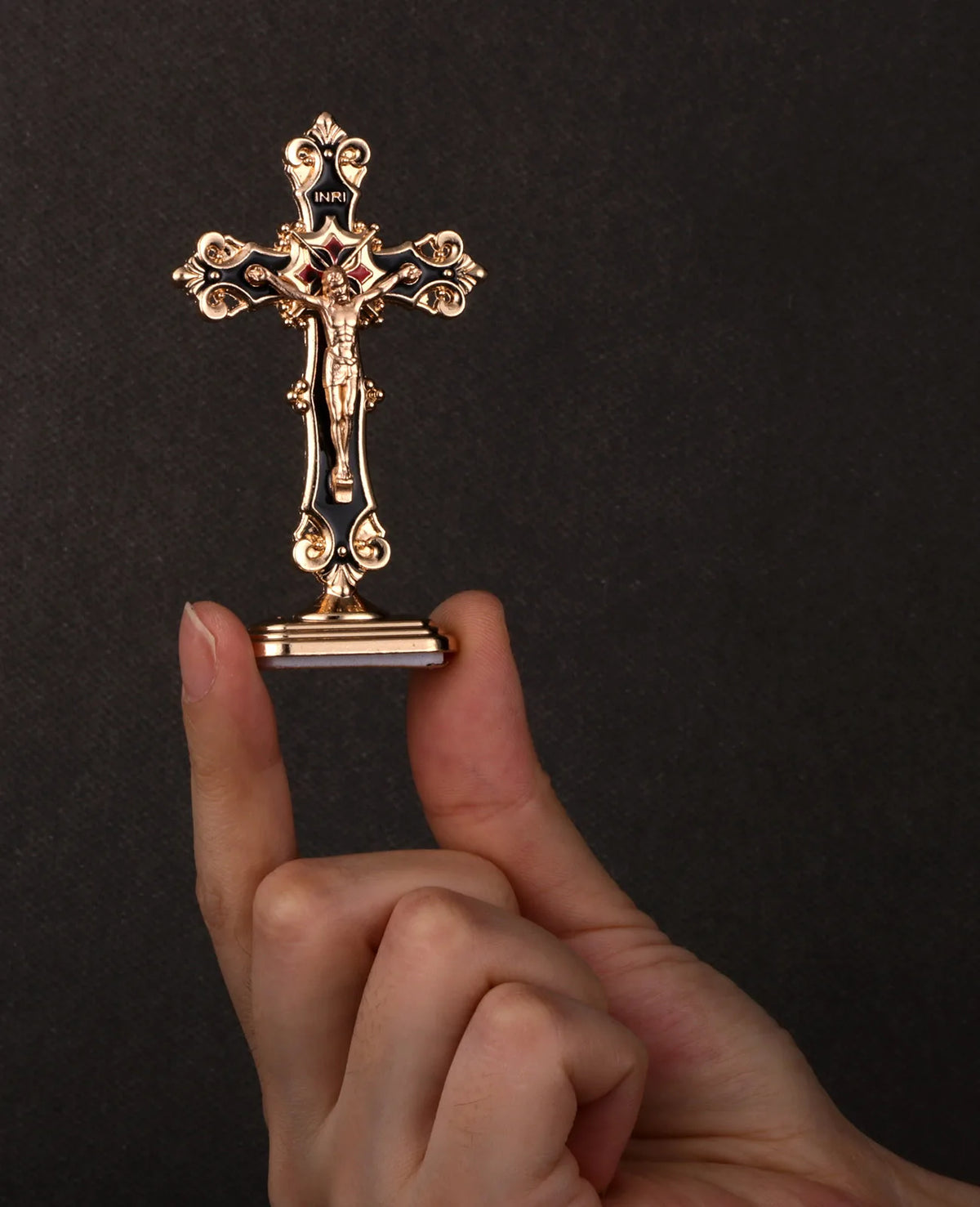 A hand holds a gold metal crucifix desk cross with a self-adhesive base against a dark background.