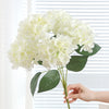 A bouquet of white artificial hydrangea silk flowers being held by a hand against a white background.