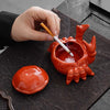 A hand is dipping a paintbrush into an orange ceramic crab ashtray with a lid on a dark slate surface.