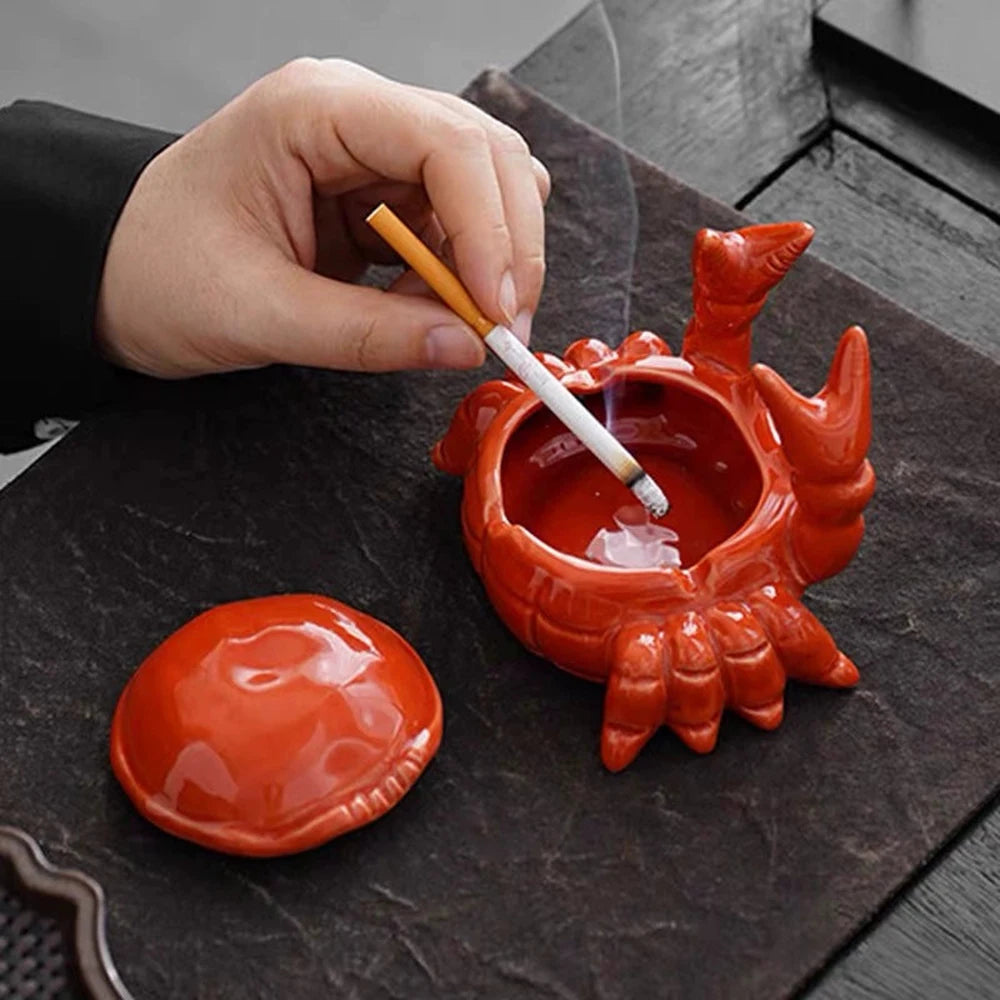 A hand is dipping a paintbrush into an orange ceramic crab ashtray with a lid on a dark slate surface.