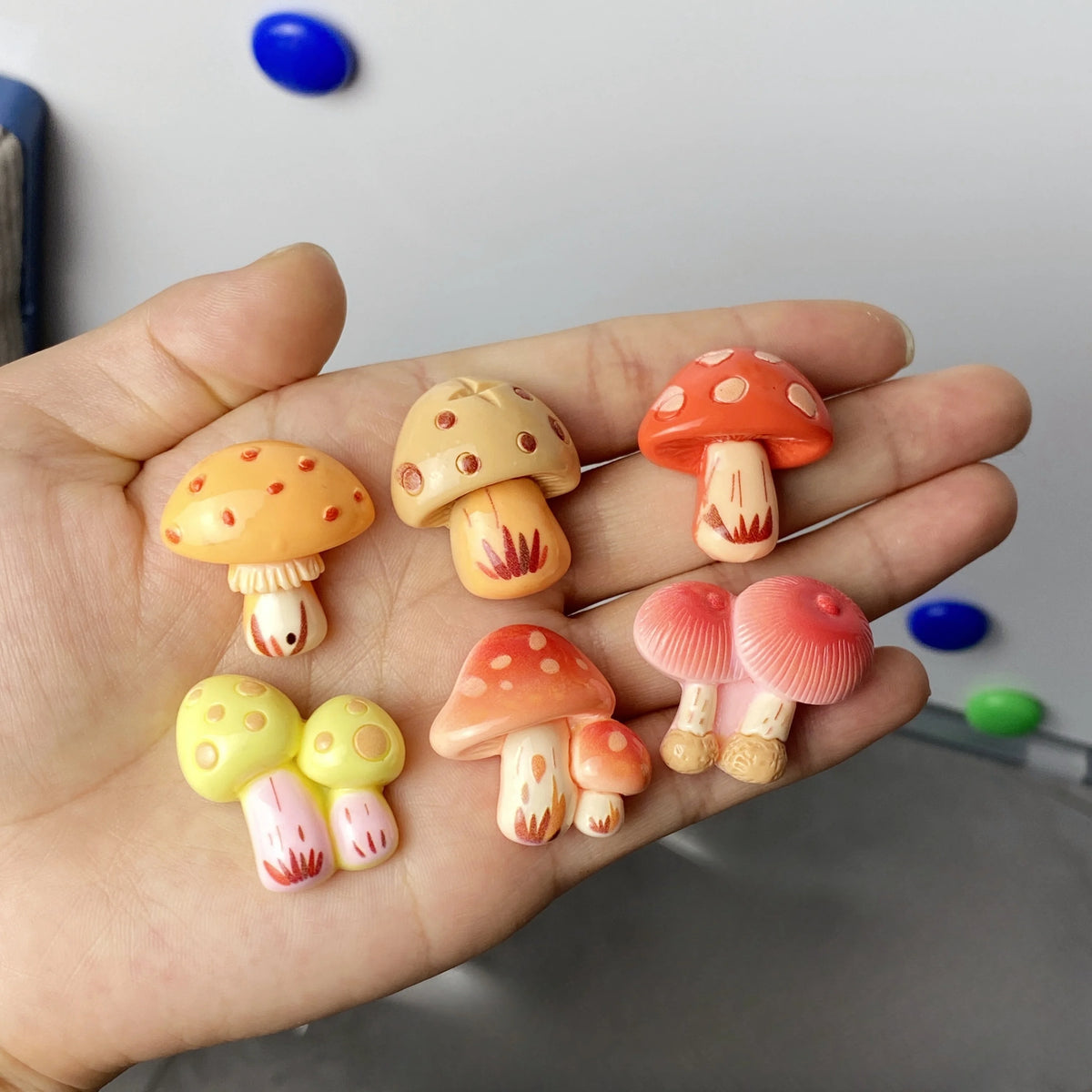 A hand holds a set of nine colorful, cute decorative mushroom fridge magnets.