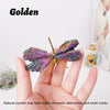 A hand holds a golden natural crystal tourmaline dragonfly figurine against a white background.